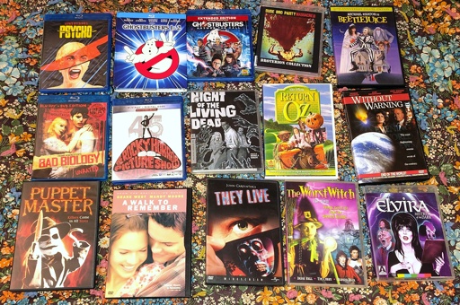 Photo of DVDs and blu-rays spread out on a blanket: the original "Psycho," "Ghostbusters" 1 and 2, "Ghostbusters" 2016, "Dude Bro Party Massacre III," "Beetlejuice," "Bad Biology," "The Rocky Horror Picture Show," the original "Night of the Living Dead," "Return to Oz," "Without Warning," "Puppet Master," "A Walk to Remember" starring Mandy Moore, "They Live," "The Worst Witch," and "Elvira, Mistress of the Dark."