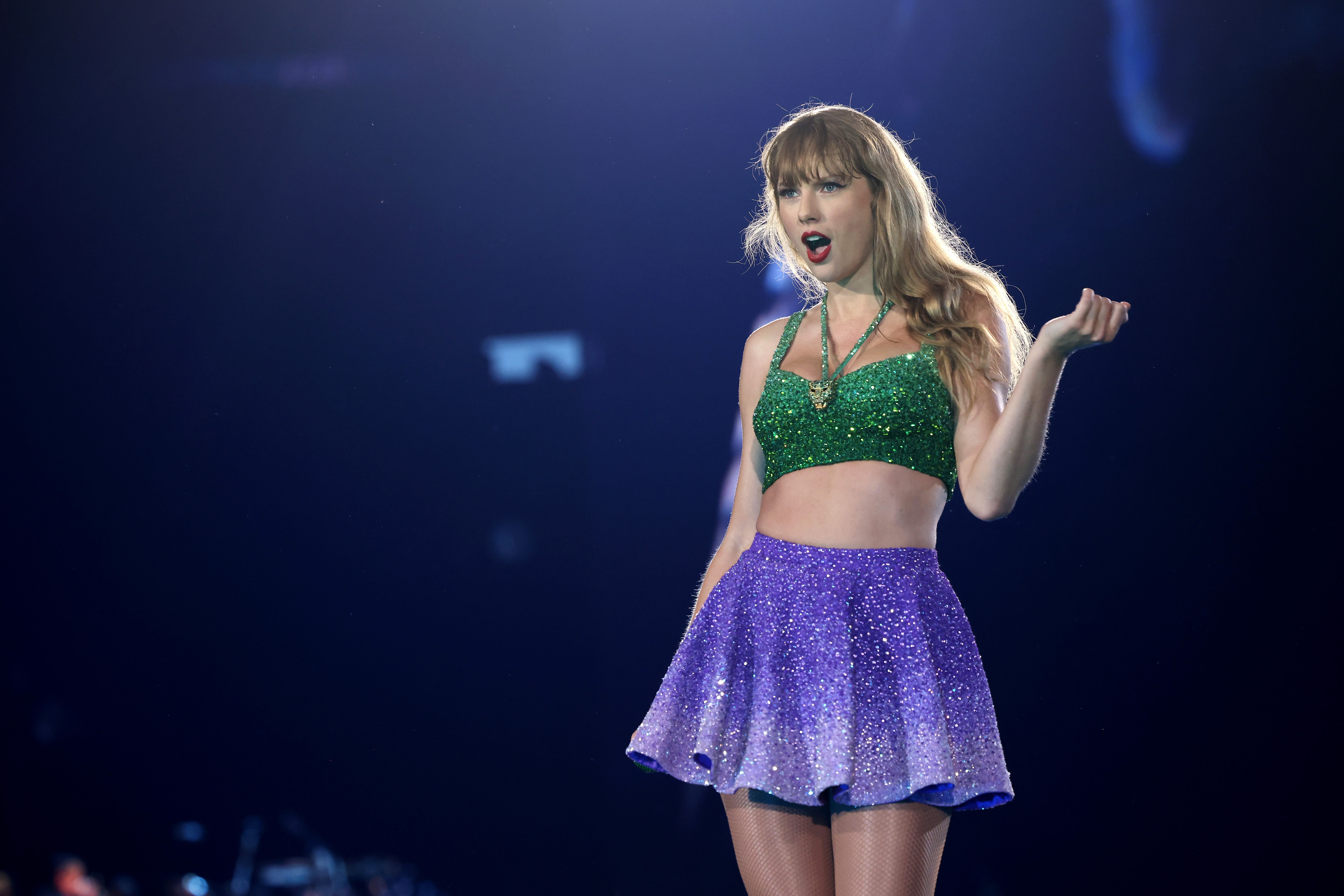 Taylor Swift, on stage at her concert, wearing a green top and purple skirt